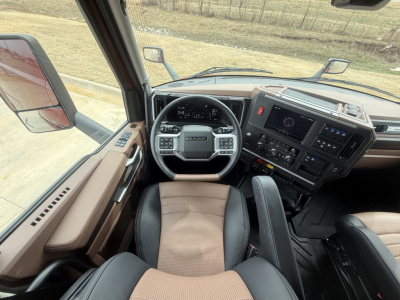 Cab interior of a Mack truck showing steering wheel with Mack badge, dashboard with multiple control panels, and tan/black seats.