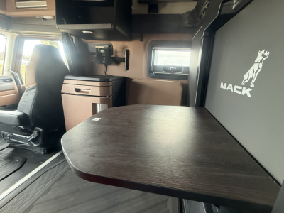 Interior of a new Mack PR64T76H truck cab with a dark wood table, storage cabinet, and a Mack logo panel visible on the right.