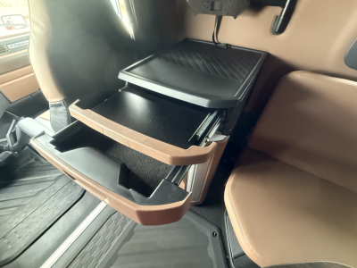 Truck cab interior showing open storage drawers and a small cooler/console in a Mack PR64T76H cabin with tan seats.
