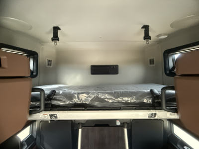 Interior sleeper cab bunk area of a new Mack PR64T76H truck showing a folded bunk with plastic cover, side cabinets, and overhead lighting.