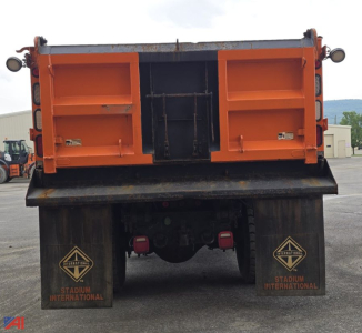 Back view of orange dump truck bed with plow, dual rear mud flaps labeled Stadium International, and mechanical chassis visible.