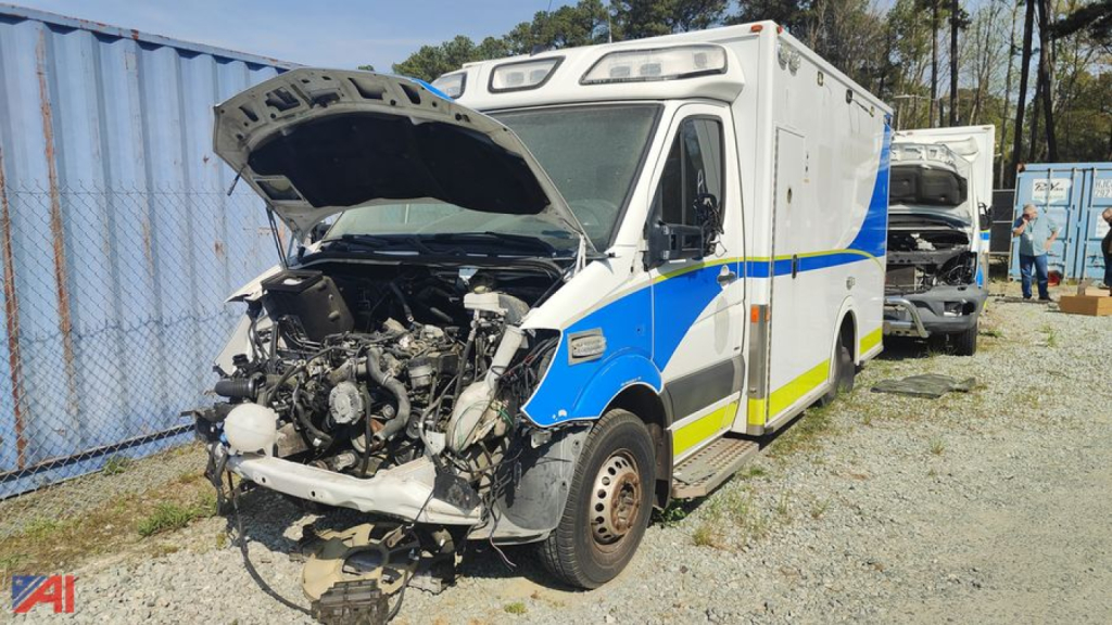 Ambulance chassis with hood open revealing engine components and front frame; body in white with blue and yellow graphics, ambulance unit parked outdoors.