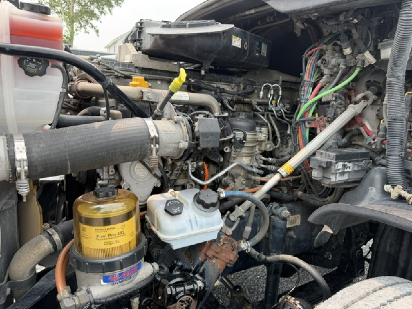 Truck engine bay showing diesel engine with yellow top filter canister, coolant reservoir, hoses, and wiring harness in a Freightliner Cascadia cab.