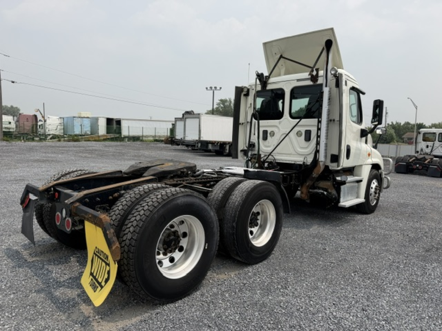 Truck tractor rear view showing fifth wheel coupling and dual rear axles with tires, Freightliner Cascadia cab, white exterior, towed in a gravel lot.