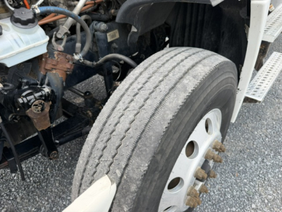 Freightliner Cascadia CA125DC front wheel well showing tire with dual tread and exposed lug nuts; engine bay and components visible on left; part of cab-side steps in view.