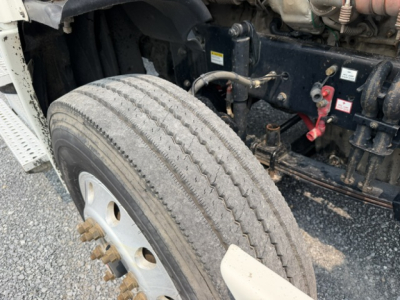 Front left wheel and chassis view of a Freightliner Cascadia CA125DC showing tire tread, wheel lug nuts, and engine bay components.