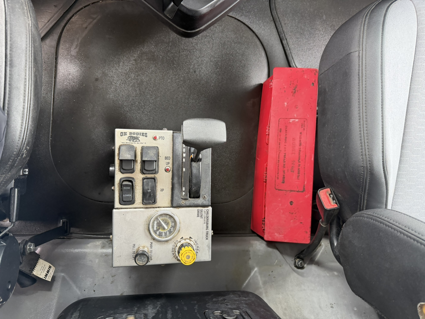 Mack GR64F dump truck interior view showing control panel with switches, gauges, and a yellow knob, plus a red rectangular box nearby.