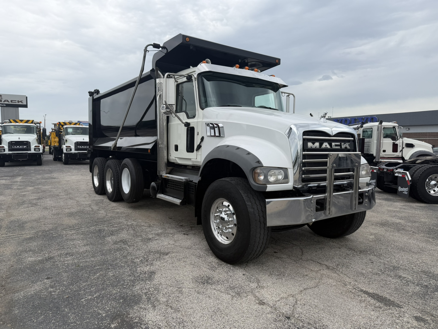 Dump truck showing front and driver side; Mack emblem on grille; white cab with black dump bed; three rear axles; chrome bumper; tires; side steps; some text on badge.