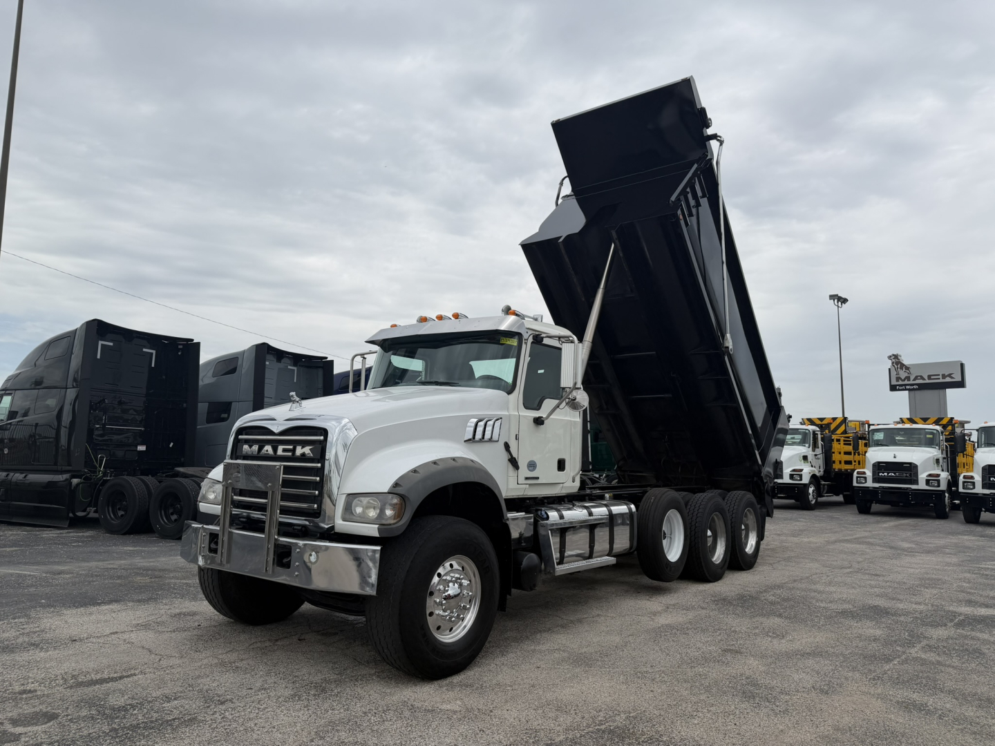Mack GR64F dump truck in a yard with the dump bed raised, front grille with Mack emblem, white cab, multiple trucks in background.