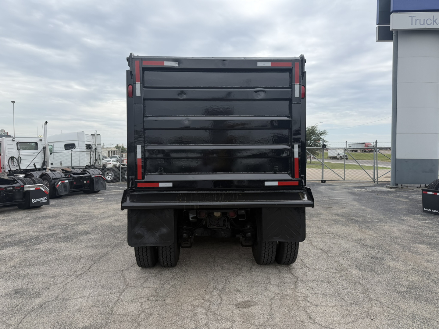 Dump truck bed rear view showing Mack GR64F with black dump body and rear double tires, tailgate closed, overcast sky.