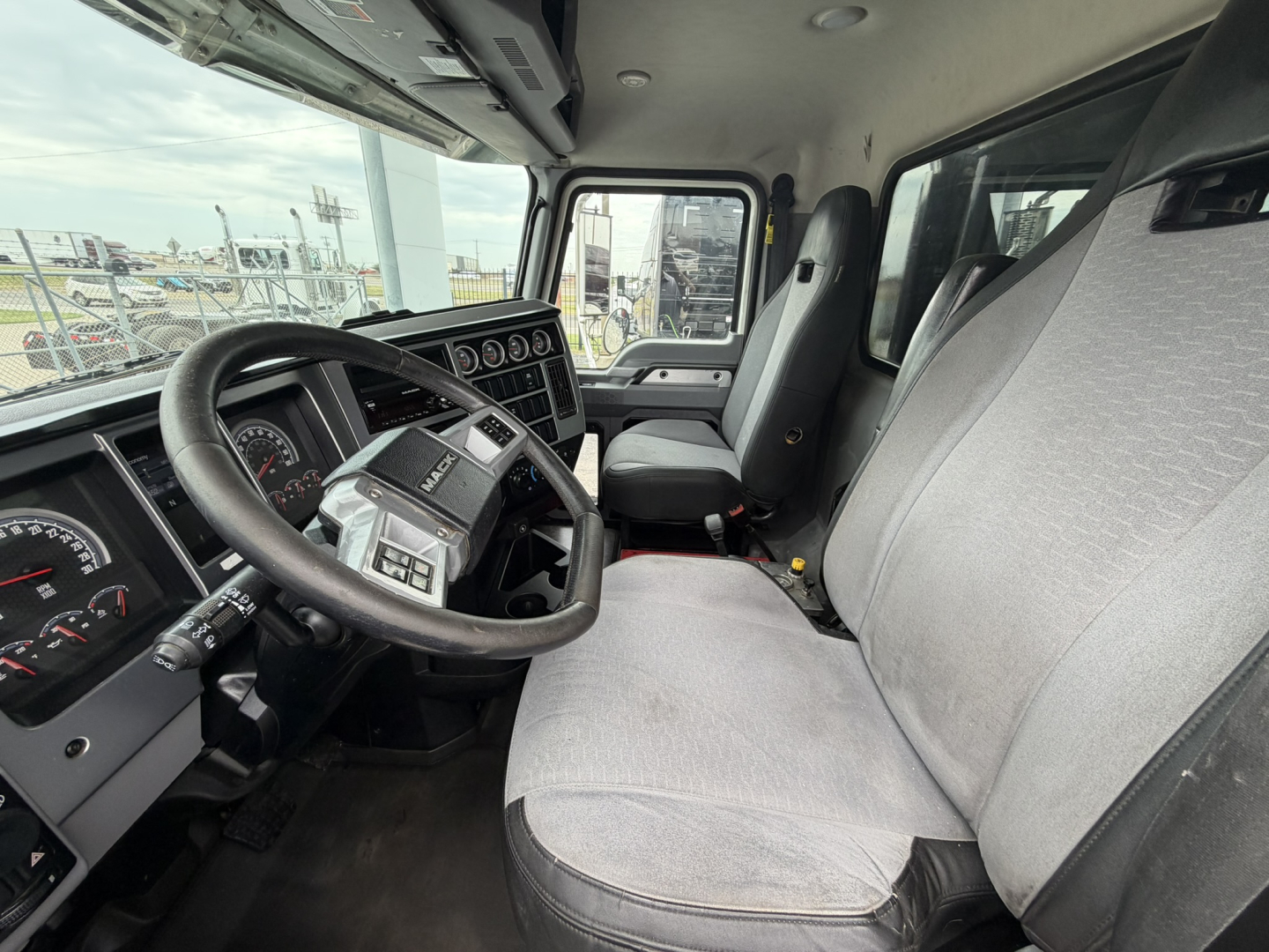 Cab interior of a Mack GR64F truck showing steering wheel, dashboard with gauges, and two front seats.