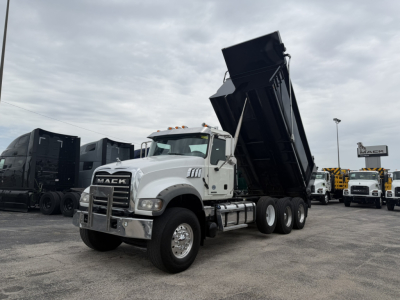 Mack GR64F dump truck in a yard with the dump bed raised, front grille with Mack emblem, white cab, multiple trucks in background.