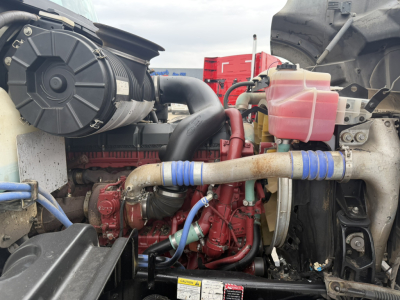 View of a Mack GR64F engine bay showing red engine block, black radiator hose, pink coolant reservoir, blue hose clamps, and exposed metal components.