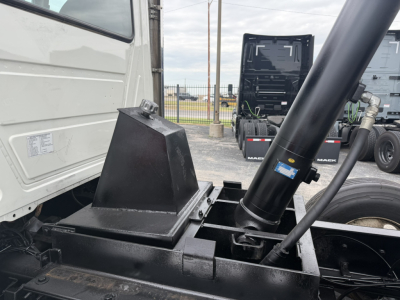Mack GR64F dump truck chassis showing hydraulic pump and mounting with black cylindrical hydraulic ram actuator and metal framework.