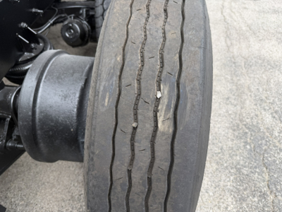Truck wheel and drive axle visible in a Mack GR64F truck showing a worn tire with tread grooves and surrounding chassis components.