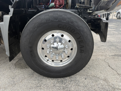 Truck front wheel area showing Goodyear tire and polished aluminum hub with multiple lug nuts; part of a Mack GR64F dump truck chassis in view.