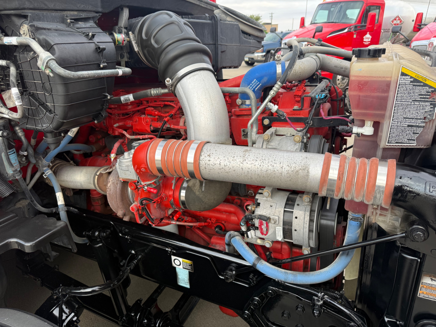 Kenworth T680 engine bay showing large turbocharged diesel engine with orange/red accents, intake hose, coolant reservoir, and various hoses.