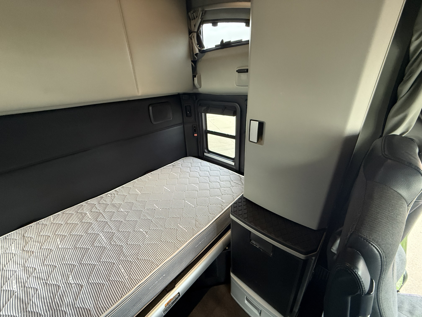Cab interior of a 2022 Kenworth T680 sleeper with bunk bed and window, showing a mattress and small cabinet near the passenger seat.