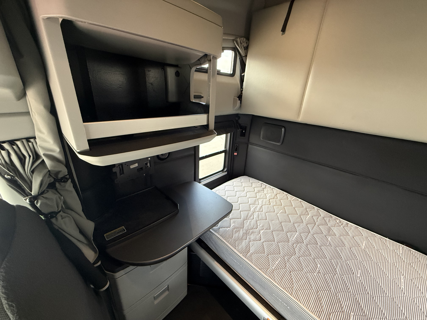 Kenworth T680 sleeper cab interior showing upper bunk area and cabin desk/bench with small shelf and drawer, light gray quilted mattress, window, and storage compartments.