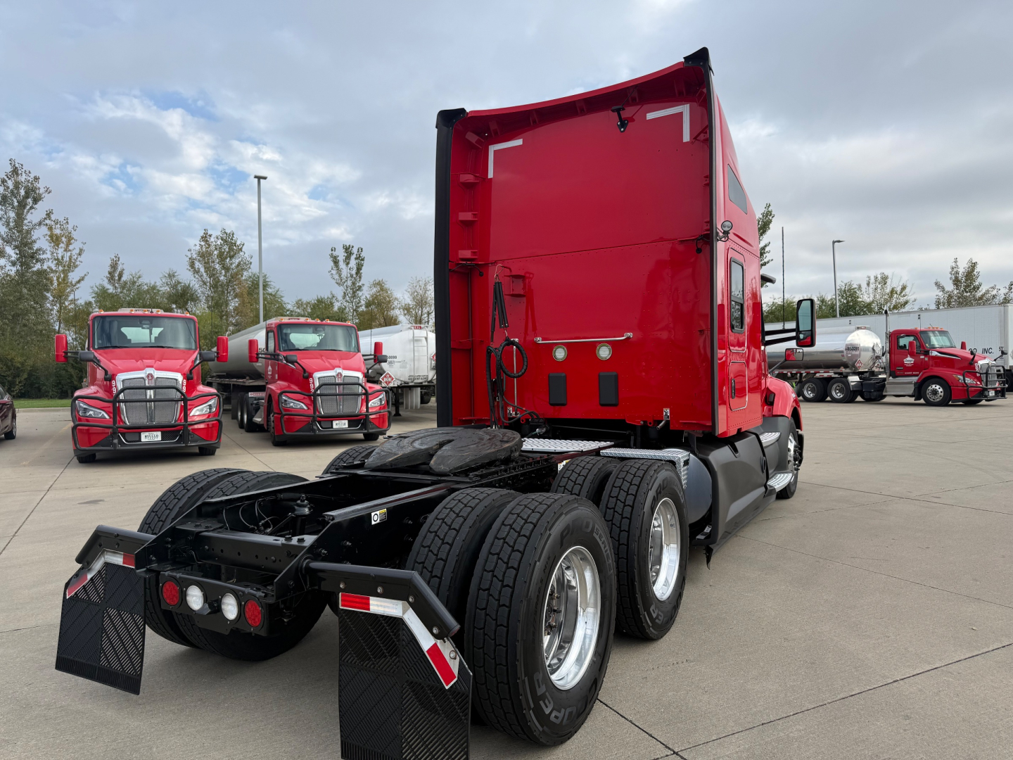 Kenworth T680 tractor with red sleeper cab, rear view showing dual rear axles and fifth wheel, parked in lot with other red trucks.