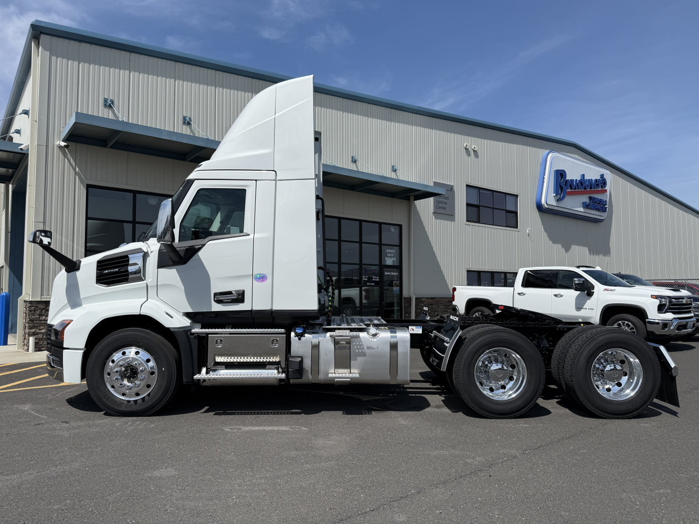 Truck cab and chassis side view of a white 2027 Mack AN64T tractor with dual rear axles, fuel tanks, and chrome wheels parked outside a dealership.