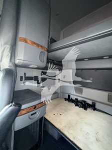 Interior sleeper cab with upper storage cabinet and flat wooden bunk surface, grey panels, and black seat belt buckles visible.