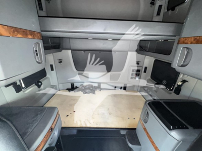 Truck cab interior showing sleeper area with beige plywood floor and overhead storage; side panels with wood veneer trim and control panels