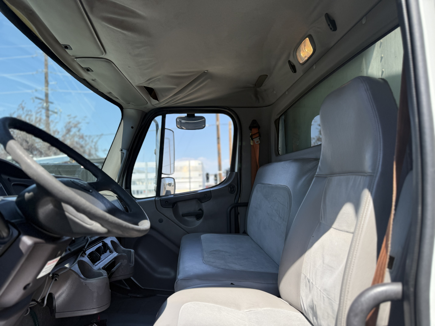 Cab interior of a Freightliner M2106 truck showing steering wheel, dashboard, and gray fabric seats with light overhead interior. No visible model text on image.