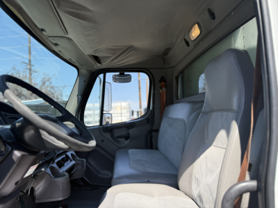 Cab interior of a Freightliner M2106 truck showing steering wheel, dashboard, and gray fabric seats with light overhead interior. No visible model text on image.