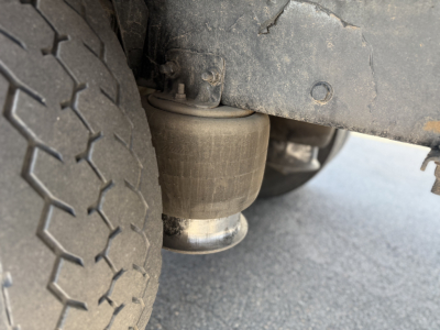 Truck undercarriage showing air suspension bag and exhaust pipe near rear tire. Visible: cylindrical air spring with metal top and bottom connections, wheel and tire tread pattern.