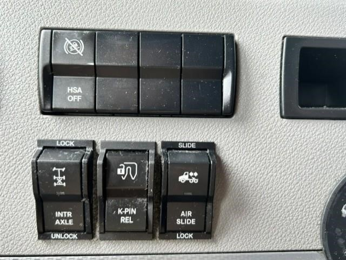 Dashboard switch panel with multiple black rocker switches labeled LOCK, INTR AXLE, K-PIN REL, AIR SLIDE; HSA OFF button; part of Freightliner Cascadia interior.