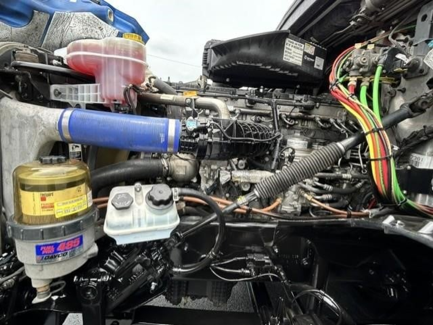 Engine bay of a Freightliner Cascadia showing hydraulic fluid reservoir, coolant reservoir, loom of colorful wires, hoses, and air intake components.