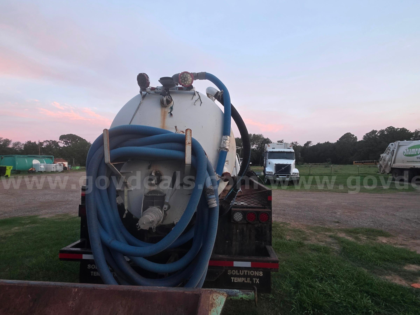 Back of a Ford F-750 vacuum truck with a large coiled blue hose on a white tank and hoses, in a gravel lot.