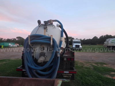 Back of a Ford F-750 vacuum truck with a large coiled blue hose on a white tank and hoses, in a gravel lot.