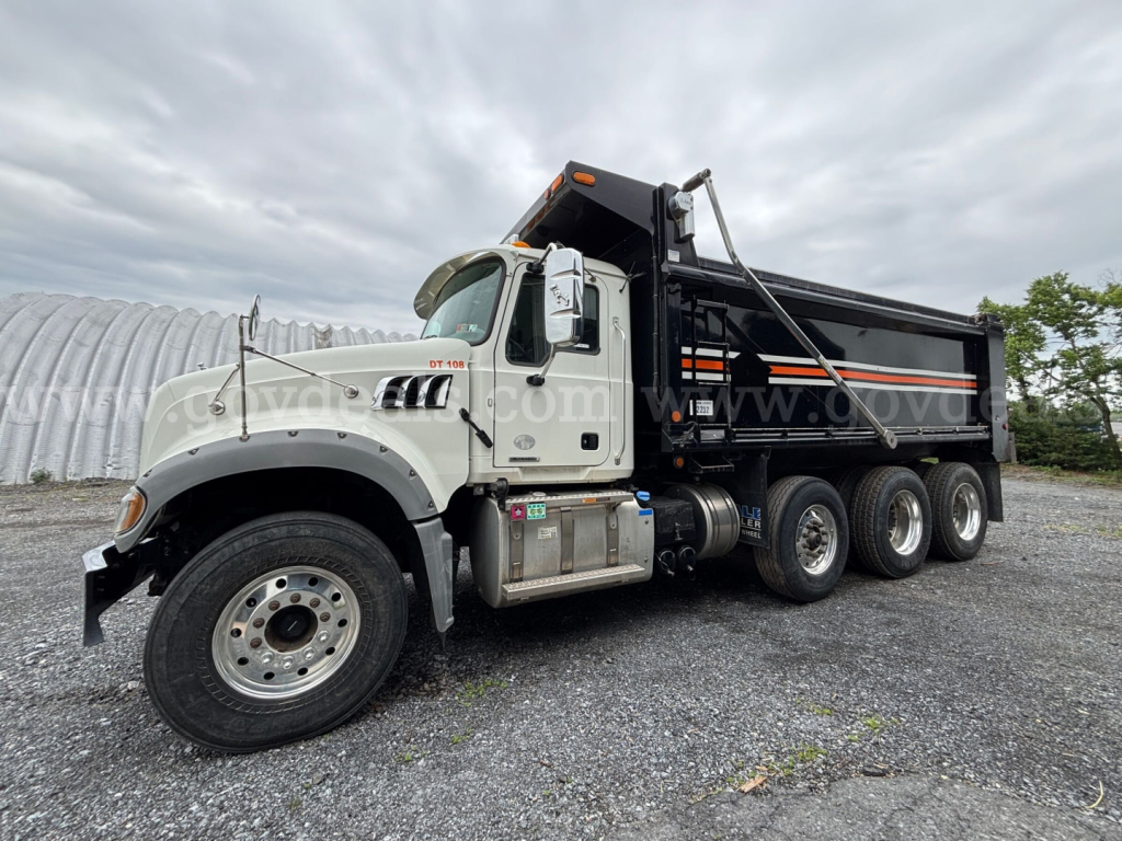 Truck cab and dump bed of a 2020 Mack Granite dump truck showing side view with three rear axles and black dump body.