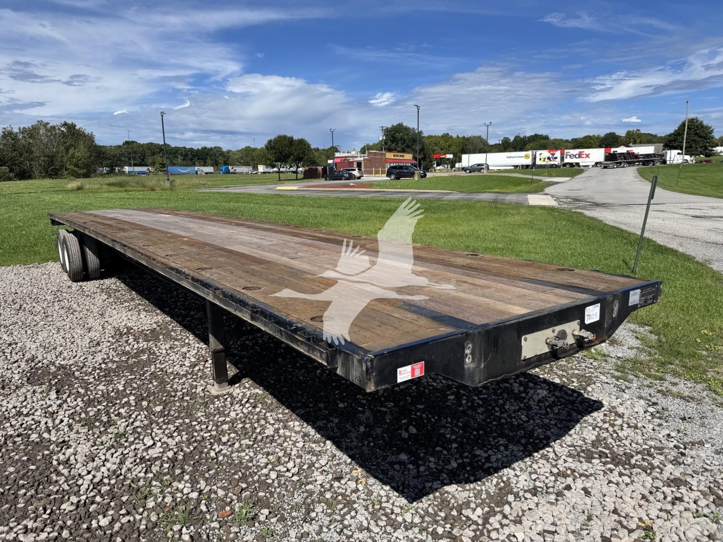 Flatbed trailer showing wooden deck and dual rear axles in an outdoor lot with clear sky.