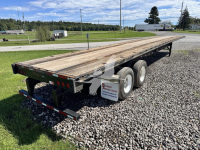 Flatbed trailer showing dual rear axles and wooden deck from rear-left perspective; stock tags and taillights visible; gravel ground.