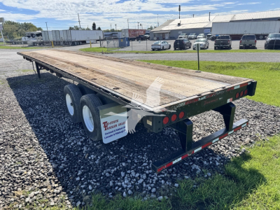 Flatbed trailer with wooden deck and dual rear axles, viewed from rear-left, showing green frame, tag lights, and mudflaps.