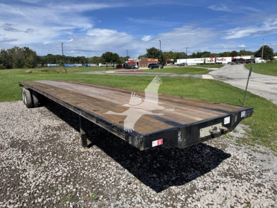 Flatbed trailer showing wooden deck and dual rear axles in an outdoor lot with clear sky.