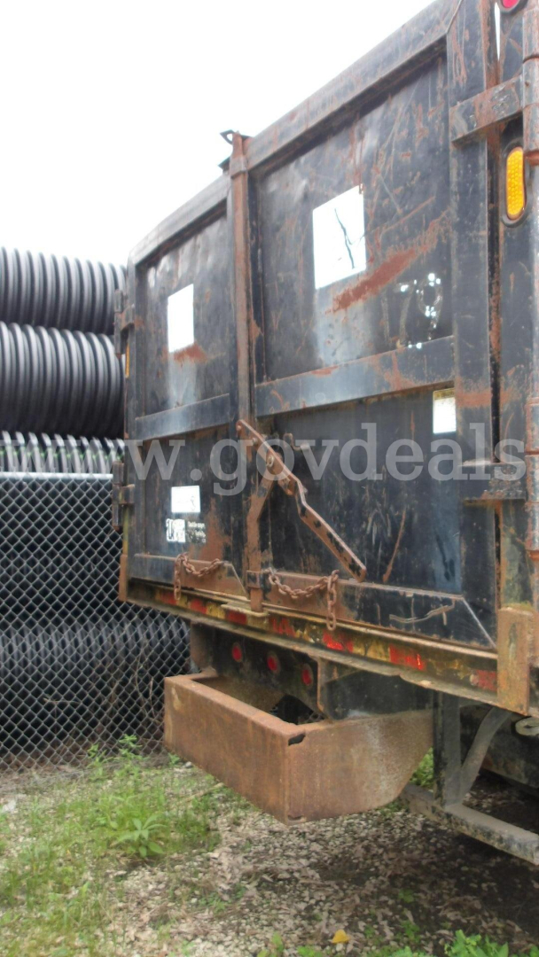 Rear of a utility truck bed with rusted metal, chain latch, and tie-down hooks; vertical taillight on the right; stacked black pipes in background.