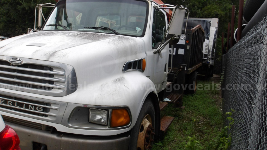 Grapple truck cab area with Sterling badge and front grille, license plate area not visible, windshield, side mirrors, and rusted wheel; appears used in outdoor lot.