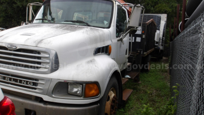 Grapple truck cab area with Sterling badge and front grille, license plate area not visible, windshield, side mirrors, and rusted wheel; appears used in outdoor lot.