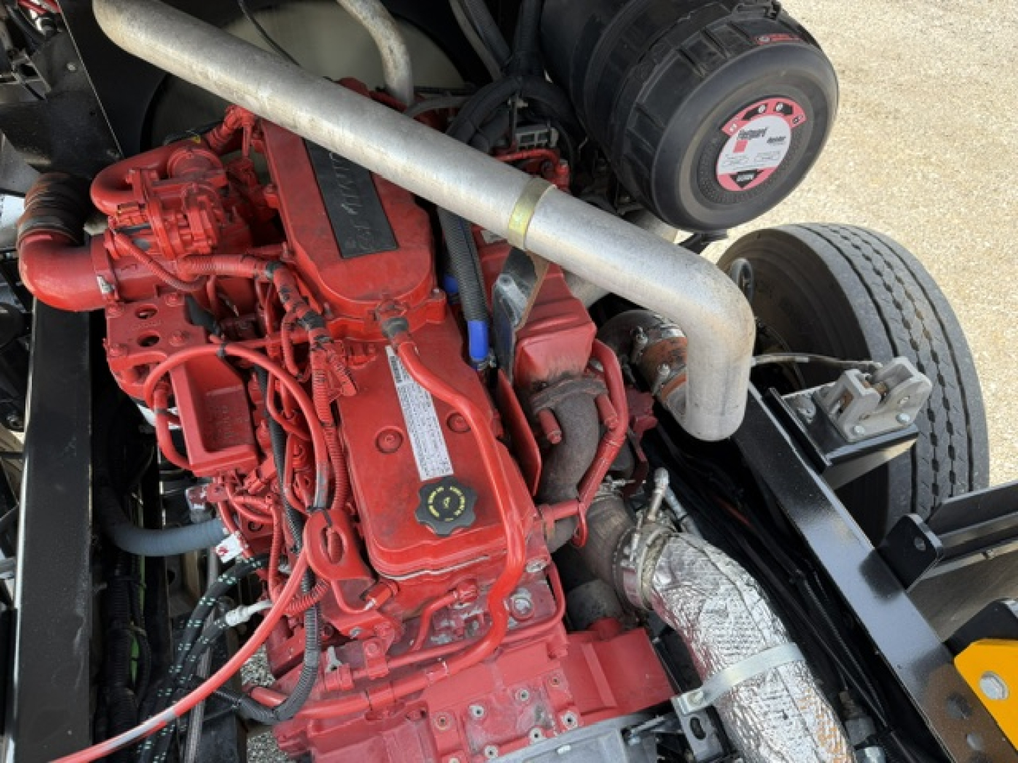 Engine bay of a red Capacity Sabre truck showing diesel engine with intake, hoses, and exhaust, plus a black circular component and metal frame.