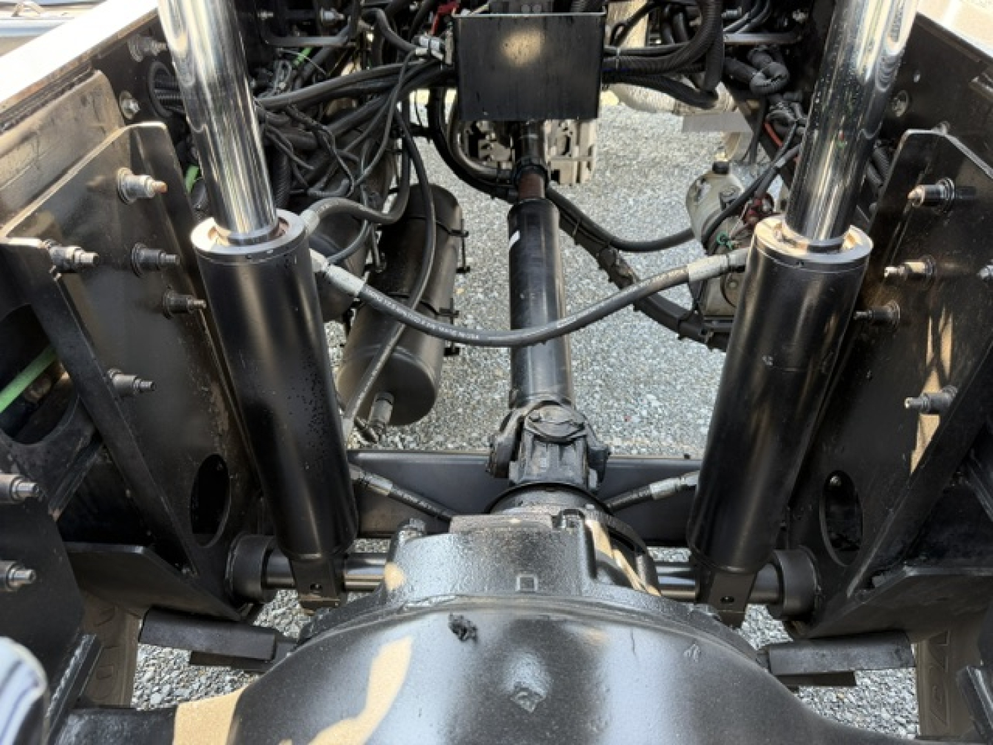 Front view of a truck chassis showing suspension components, drive shaft, and hydraulic lines; hydraulic cylinders on each side, axles, and universal joints visible.