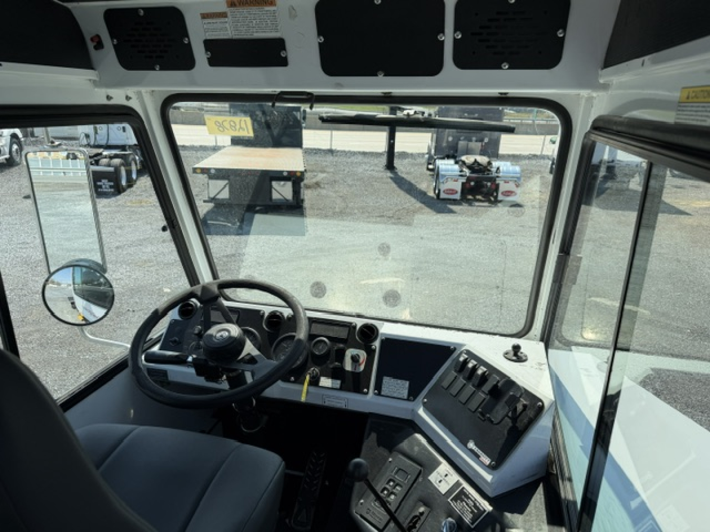 Cab interior of a truck showing steering wheel, dashboard with gauges and switches, large windshield, side windows, and overhead panels.