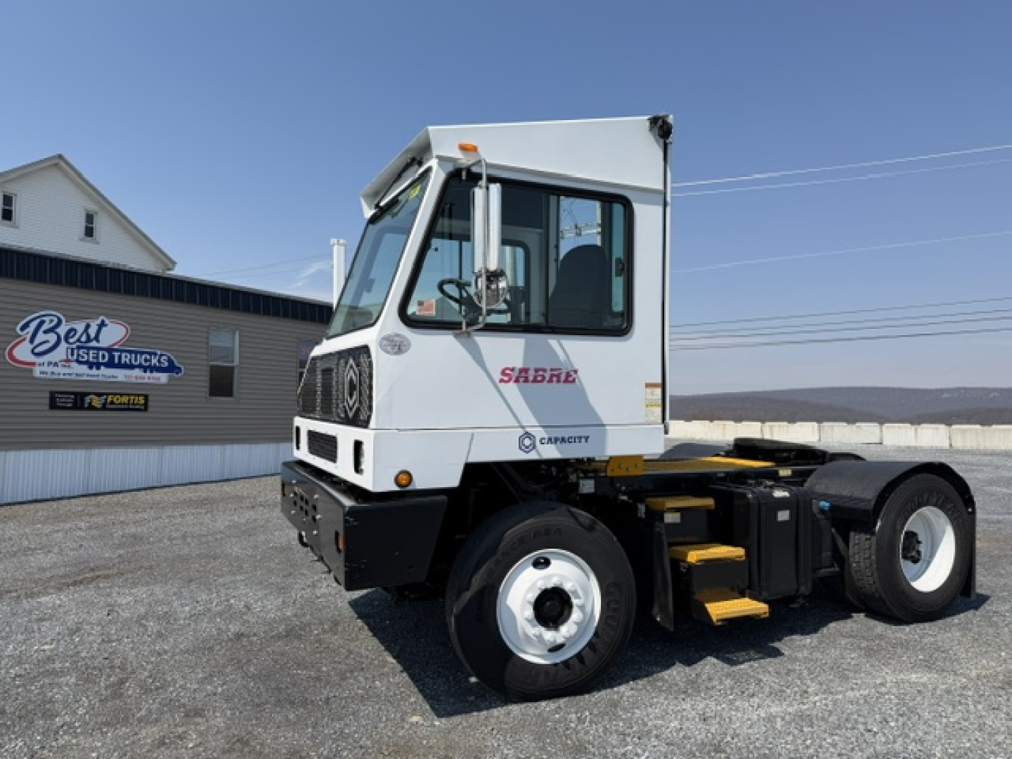 Truck cab with 'SABRE' logo and Capacity brand on side; cab window, door, and step; wheel and chassis visible; text 'CAPACITY' near rear; yard in background.