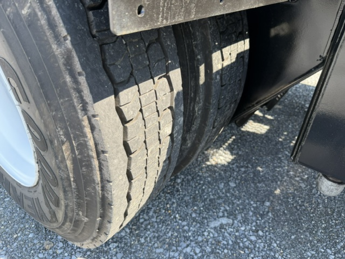 Truck wheel/front tire view showing dual rear tires and wheel well. Visible tread and sidewall text, part of chassis and undercarriage.