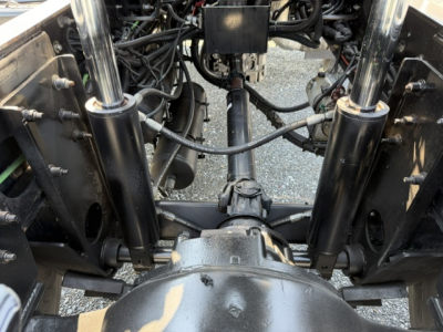 Front view of a truck chassis showing suspension components, drive shaft, and hydraulic lines; hydraulic cylinders on each side, axles, and universal joints visible.
