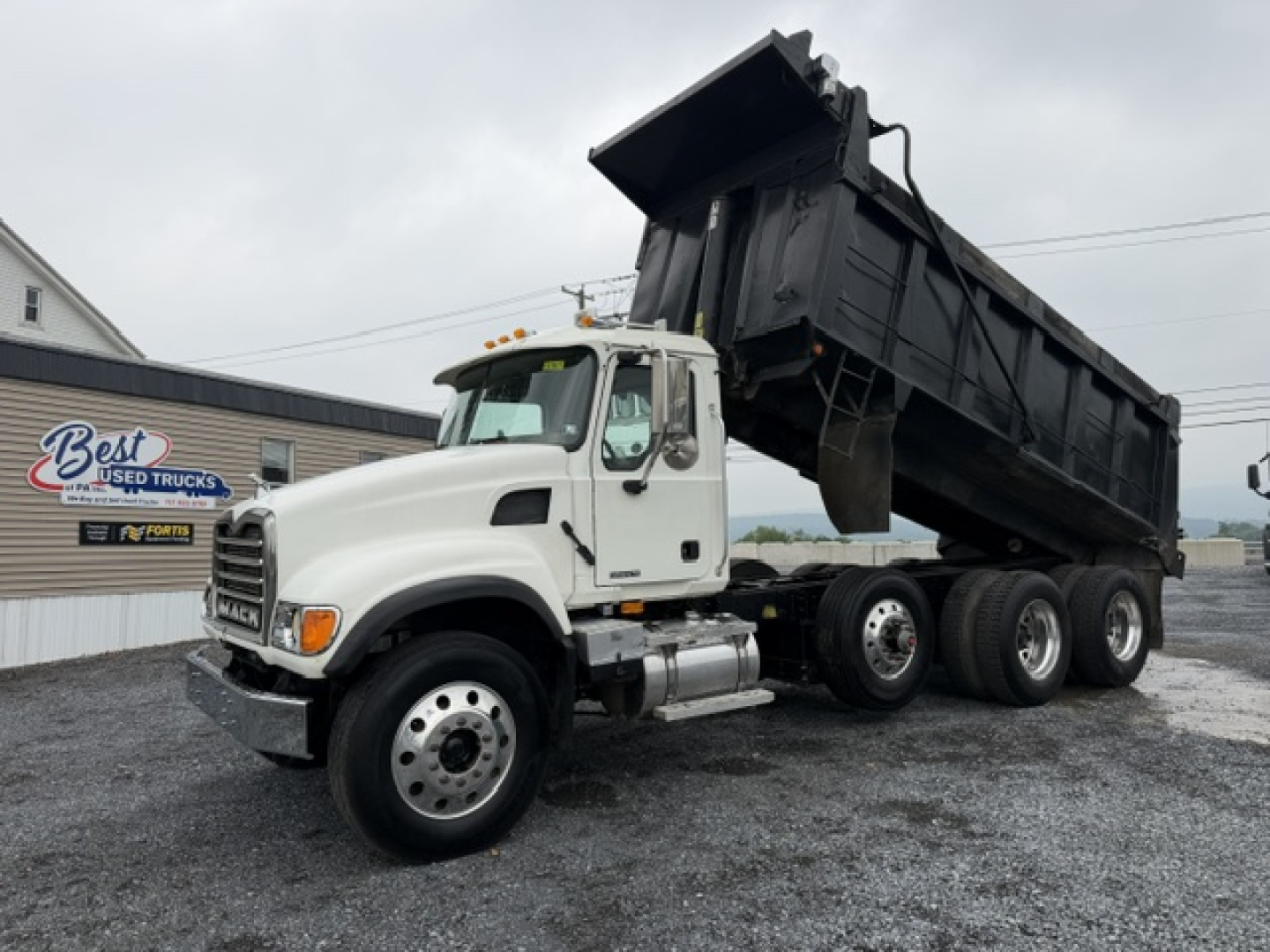 Dump truck with raised black dump bed on a white Mack CV713 tractor, side view showing dual rear axles and chrome fuel tanks.