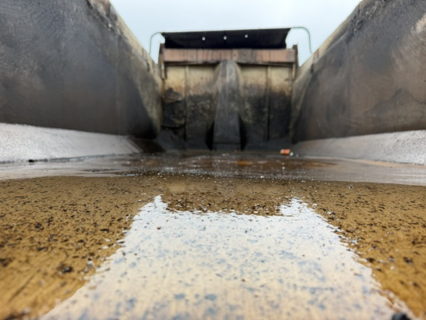 Dump truck bed interior with dirt and a puddle, viewed from near the front edge toward the tailgate.